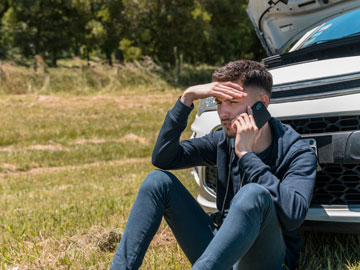 Junger Mann sitzt vor dem Auto und teilt seinen Schaden mit
